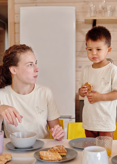 A woman and a young boy wearing matching cream-colored t-shirts with a small bird graphic, sitting and standing at a breakfast table in a kitchen setting.